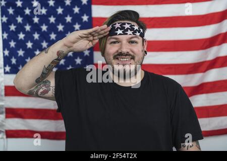 Homme caucasien barbu avec un bandana souriant tout en donnant un Saluez devant le drapeau américain Banque D'Images