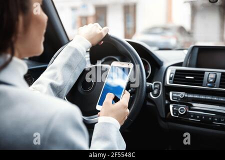 Photo courte des mains de la femme tenant le volant et le smartphone. Femme d'affaires utilisant l'application de téléphone mobile pendant la conduite, en vérifiant l'itinéraire sur son bureau de chemin Banque D'Images