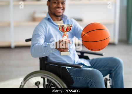 Joyeux sportif noir en fauteuil roulant, montrant le basket-ball et le trophée, heureux de sa victoire à la maison, foyer sélectif Banque D'Images