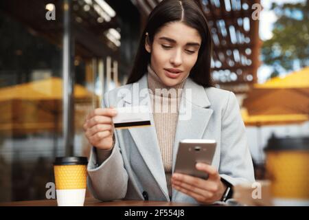 Jeune femme payant en ligne, en utilisant une carte de crédit et un téléphone mobile tout en étant assise dans un café, transférer de l'argent par smartphone ou faire des achats sur Internet Banque D'Images