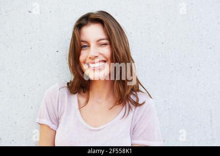 Portrait en gros plan d'une jeune femme heureuse souriant et en se clin d'œil contre le mur gris Banque D'Images