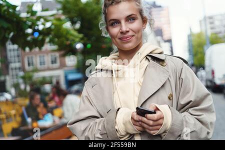 Gros plan de la belle jeune femme debout dans la rue avec téléphone mobile et sourire heureux. Fille attendant petit ami près du café. Messages féminins Banque D'Images