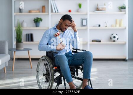 Portrait d'un triste sportif noir avec trophée assis en fauteuil roulant, se sentant déprimé au-dessus de sa blessure à la maison Banque D'Images