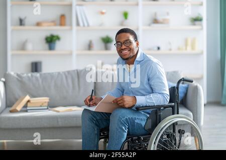 Joyeux homme noir handicapé en fauteuil roulant prenant des notes, regardant l'appareil photo et souriant à la maison, espace de copie Banque D'Images