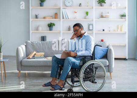 Homme noir handicapé dans un carnet de lecture en fauteuil roulant, travaillant ou étudiant à la maison pendant l'épidémie de Covid-19 Banque D'Images