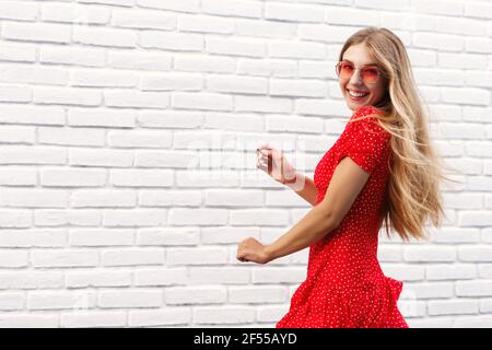 Concept de vacances d'été. Bonne fille blonde dansant dans les rues en robe rouge et lunettes de soleil, souriant à l'appareil photo, appréciant les vacances d'été Banque D'Images