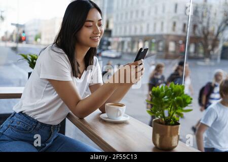 Belle fille asiatique discutant sur téléphone portable tout en étant assise dans un café de la ville près de la fenêtre, boire un café et utiliser un smartphone, souriant comme écran de lecture Banque D'Images