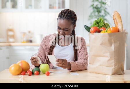 Des produits d'épicerie chers. Une femme de ménage africaine choquée assise à la table dans la cuisine, en vérifiant les factures Banque D'Images