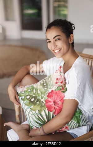 Notion de tendresse, d'émotions et de confort. Tir vertical mignonne hispanique brune femme en t-shirt blanc, jambes assis croisées dans confortable rotin Banque D'Images