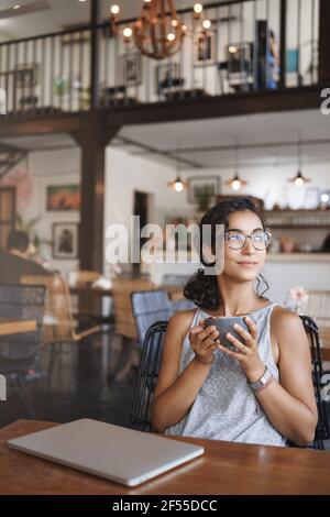 Prise de vue verticale réussie femme attirante freelance numérique nomade profiter de l'été vue sur la nature urbaine s'asseoir près d'un ordinateur portable fermé table de café tasse à café Banque D'Images