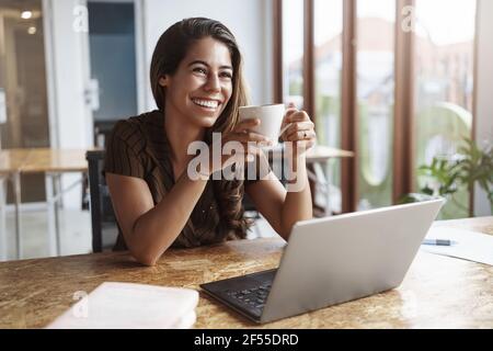 Le travail va vite, bonne équipe. Charmante femme d'affaires insouciante tenir tasse de café rire regarder collègue souriant plaisantant travail ordinateur portable étudier Banque D'Images