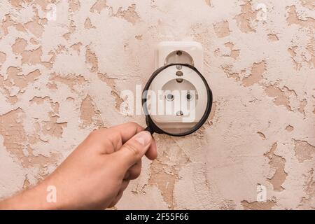 La main de l'homme tient une loupe et examine une double douille blanche sur un mur abstrait moderne dans une pièce rapprochée. Banque D'Images