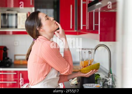 Belle femme léchant le doigt tout en préparant des fruits smoothie dans la cuisine à la maison Banque D'Images