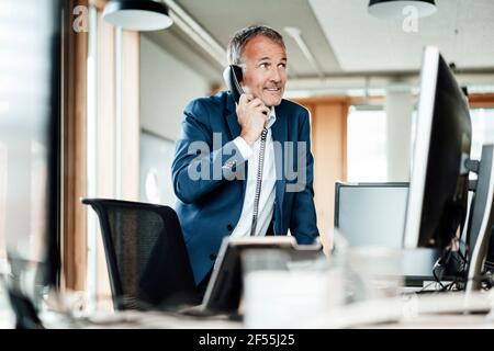 Homme d'affaires souriant regardant loin en parlant au téléphone à l'avant de l'ordinateur au bureau Banque D'Images