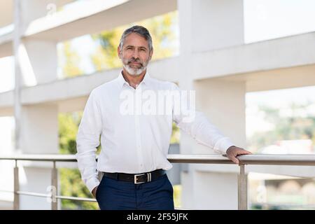 Homme d'affaires confiant et mature debout avec les mains dans les poches par garde-corps sur la terrasse du bureau Banque D'Images