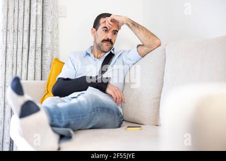 Homme inquiet portant une élingue de bras assis avec la tête à la main sur le canapé à la maison Banque D'Images