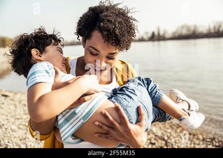 Mère souriante prenant son fils en se tenant au bord du lac Banque D'Images