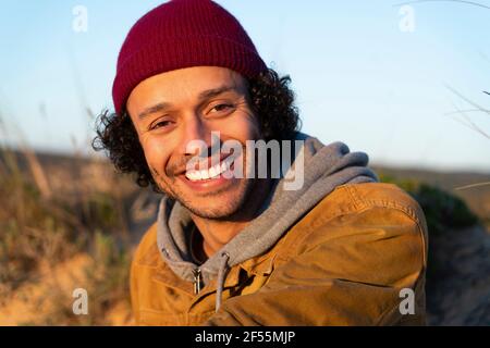 Jeune homme portant un chapeau en tricot souriant tout en étant assis à l'extérieur Banque D'Images