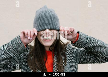 Femme souriante couvrant les yeux à travers un chapeau tricoté contre le mur Banque D'Images