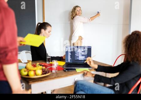 Femme d'affaires souriante discutant avec ses collègues de la stratégie d'entreprise sur le tableau blanc pendant la réunion au bureau Banque D'Images