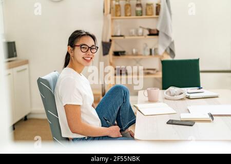 Jeune femme souriante assise près d'une table à la maison Banque D'Images