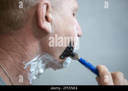 Un homme européen âgé avec de la mousse sur son visage se rase à l'aide d'une machine. La vie de personnes réelles. Banque D'Images