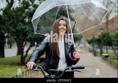 Femme souriante avec vélo électrique tenant un parapluie dans la rue en ville Banque D'Images