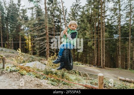 Garçon assis sur une balançoire à pneus dans la forêt pendant les vacances à Salzburger Land, Autriche Banque D'Images