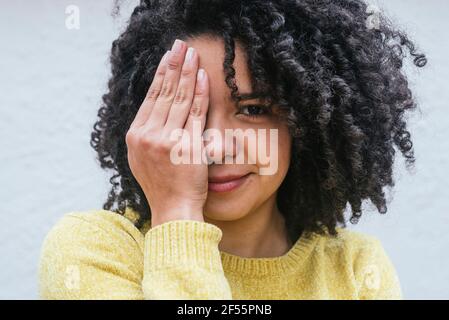 Femme aux cheveux bouclés couvrant l'œil contre un mur blanc Banque D'Images