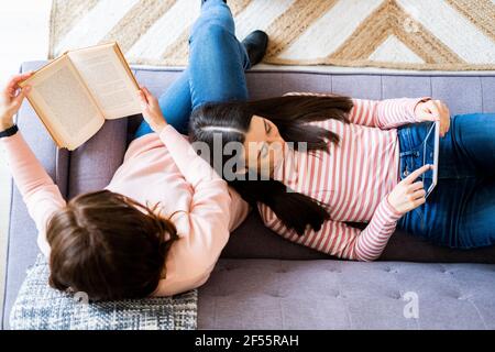 Mère lisant un livre et fille utilisant une tablette numérique pendant qu'elle mentait sur le canapé dans la salle de séjour Banque D'Images