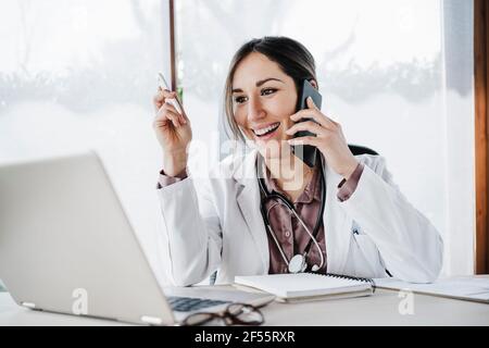 Femme souriante médecin regardant un ordinateur portable tout en parlant sur mobile téléphone à l'hôpital Banque D'Images