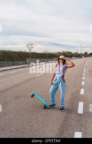 Femme souriante en chapeau debout avec planche à roulettes sur la route Banque D'Images