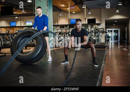Des amis d'athlète de sexe masculin s'entraînant avec une corde et un pneu en salle de sport Banque D'Images
