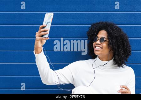 Adolescent souriant portant des écouteurs intra-auriculaires pour faire passer le selfie en mobile téléphone assis contre un mur bleu Banque D'Images