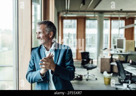 Homme d'affaires souriant avec les mains jointes regardant à travers la fenêtre dedans bureau Banque D'Images