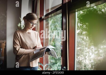 La femme aime passer du temps avec la lecture. Photo d'intérieur d'une jolie femme créative et élégante dans une tenue et des lunettes tendance, avec un magazine et des pages tournantes Banque D'Images