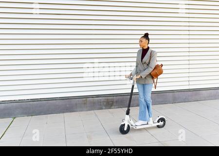 Jeune femme d'affaires avec sac à cheval électrique push scooter contre le mur rayé blanc Banque D'Images