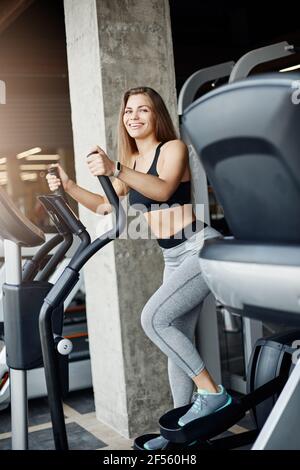 Jeune belle athlète féminine utilisant l'entraînement elliptique pour réchauffer ses muscles. Courir dans la salle de sport un matin d'hiver ensoleillé. Banque D'Images