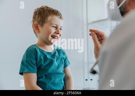 Garçon souriant en ayant un écouvillon nasal assis par un médecin à clinique Banque D'Images