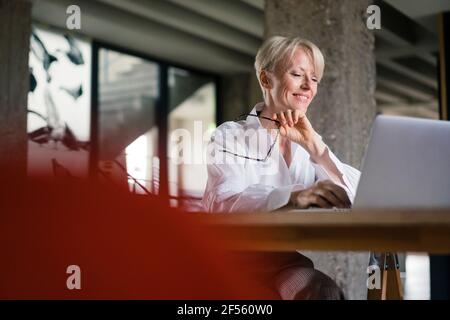 Travailleur indépendant souriant tenant des lunettes tout en utilisant un ordinateur portable au bureau au bureau à domicile Banque D'Images