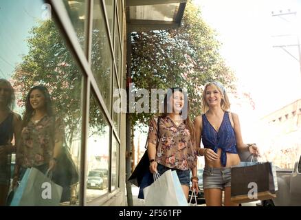 Des femmes souriantes portant des sacs de shopping tout en marchant près d'une fenêtre en verre en ville Banque D'Images