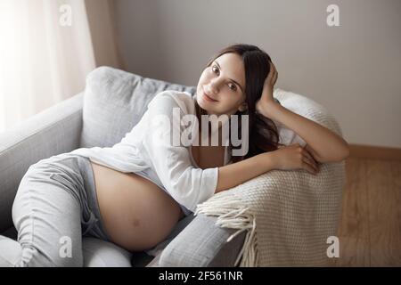 Bébé veut que vous toudiez le ventre. Portrait d'une femme européenne enceinte heureuse et détendue dans une tenue confortable et confortable, assise dans un fauteuil, penchée sur la paume et Banque D'Images