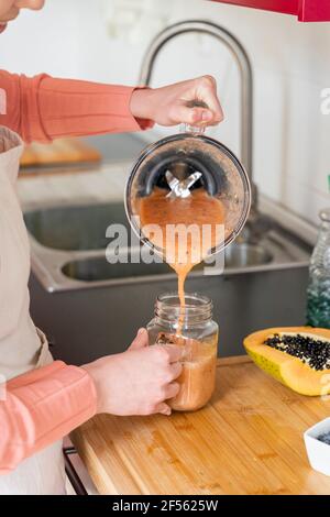 Jeune femme versant du smoothie dans un pot de maçon dans la cuisine à accueil Banque D'Images