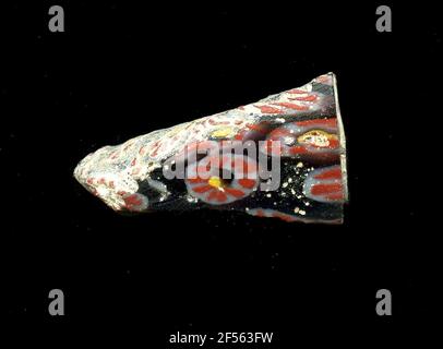 Fragment d'un vaisseau. Une faible courbure du fragment de verre permet l'affectation à un vaisseau, éventuellement à une coque. Sur fond noir, plusieurs fleurs sont présentées dans la technologie millefiori. Autour d'un point jaune se trouve une bande blanche, qui rayonne plusieurs traits rouges sont les pétales. Pour la production, des verres de différentes couleurs ont été formés en tubes et en tiges. Elles étaient comprimées et chauffées, ce qui entraînait une longue barre. Le verre coupé en tranches de ​​now avait des motifs amorphes ou floraux différents. Par la suite, plusieurs de ces plaquettes ont été repliées ensemble, réchauffées à nouveau et pressées en forme vasculaire. Burry pour Banque D'Images