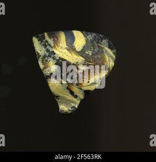 Fragment d'un vaisseau. Une faible courbure du fragment de verre permet l'affectation à un vaisseau, éventuellement à une coque. Dans le verre brun, plusieurs bandes jaunâtres et grises peuvent être reconnues irrégulièrement. D'autre part, un point rouge. Pour la production, des verres de différentes couleurs en tubes et en tiges ont été formés. Elles étaient comprimées et chauffées, ce qui entraînait une longue barre. Le verre coupé en tranches de ​​now avait des motifs amorphes ou floraux différents. Par la suite, plusieurs de ces plaquettes ont été repliées ensemble, réchauffées à nouveau et pressées en forme vasculaire. Burry pour le processus de fabrication élaboré et son fr Banque D'Images