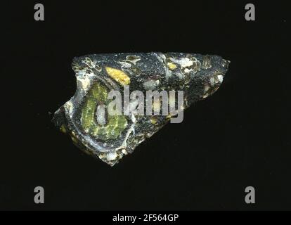 Fragment d'un vaisseau. Une faible courbure du fragment de verre permet l'affectation à un vaisseau. La raison du verre est vert olive foncé. Ceux-ci sont de voir des fleurs dans la technologie Millefiori. Autour d'un point blanc, un ruban vert se présente sous forme de pétales. Sur le bord de la fracture se trouvent les points jaunes, qui sont disposés circulaire et aussi les pétales de pollen. Pour la production, des verres de différentes couleurs ont été moulés dans des tubes et des tiges. Elles étaient comprimées et chauffées, ce qui entraînait une longue barre. Le verre coupé en tranches de ​​now avait des motifs amorphes ou floraux différents. Par la suite, plusieurs de ces plaquettes ont été fusionnées, réchauffées à nouveau Banque D'Images