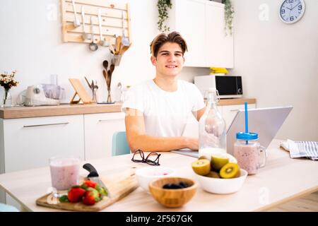 Jeune homme avec ordinateur portable assis à une table dans la cuisine Banque D'Images