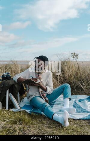 Jeune homme avec journal assis sur une couverture à la plage Banque D'Images