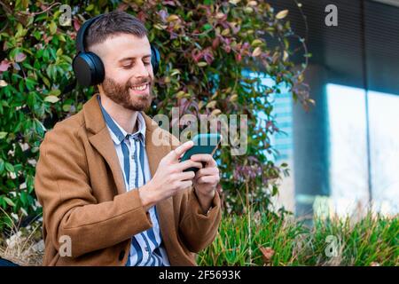 Homme d'affaires souriant portant un casque à l'aide d'un smartphone en étant assis à l'extérieur Banque D'Images