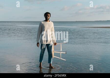 Jeune homme avec tabouret blanc debout à la plage contre le ciel Banque D'Images